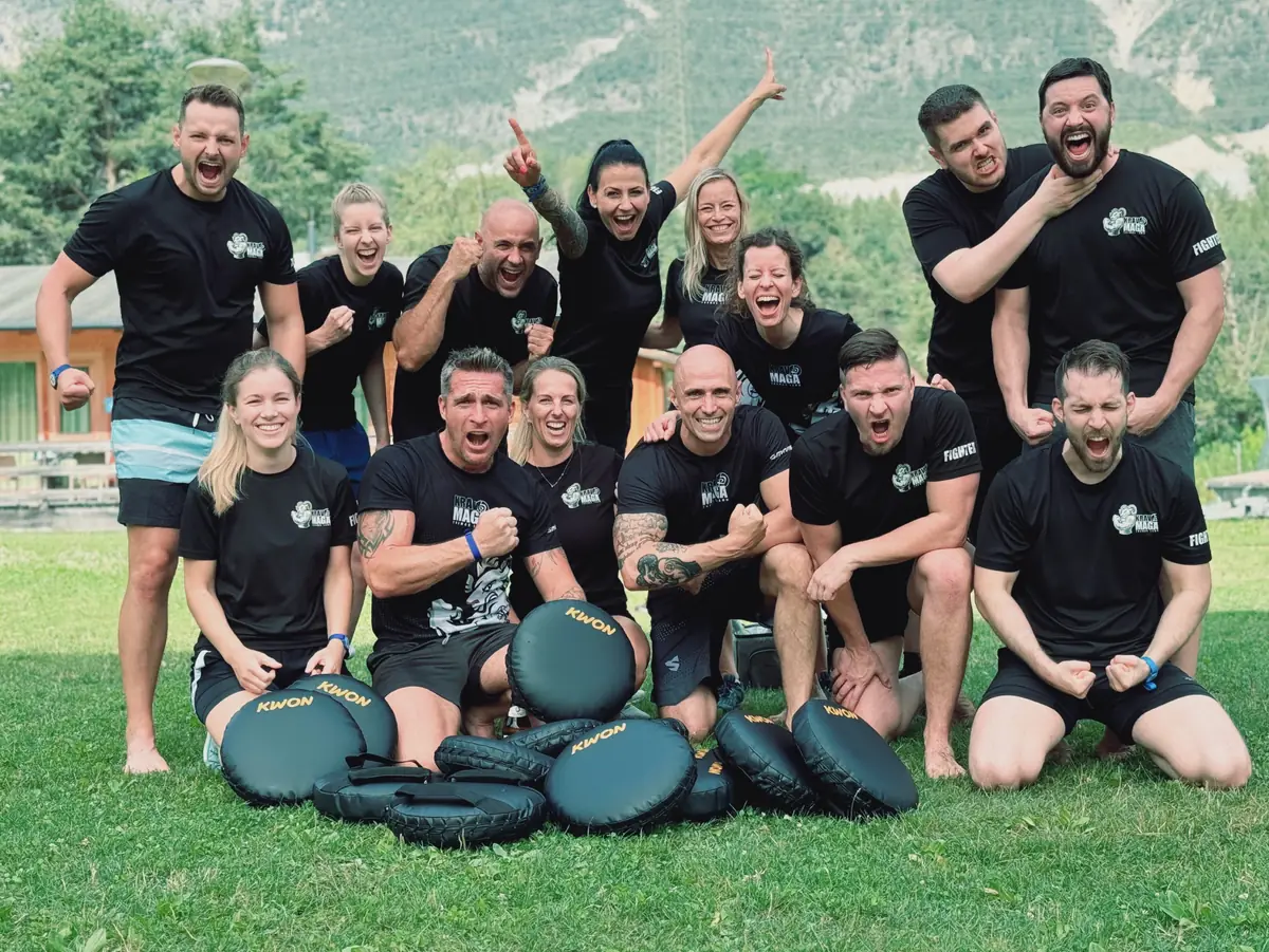 Die Gruppe in schwarzen Team-Shirts jubelt auf einer Wiese, vor ihr liegen Schlagpolster, dahinter Berge