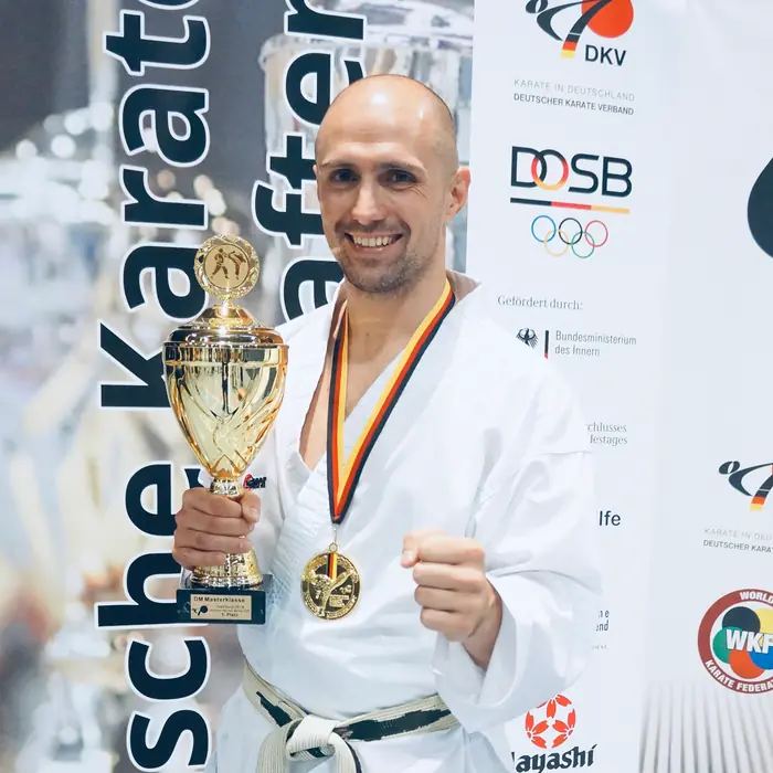 Thomas Lamm im Karateanzug mit Pokal und Goldmedaille vor einer Sponsorenwand