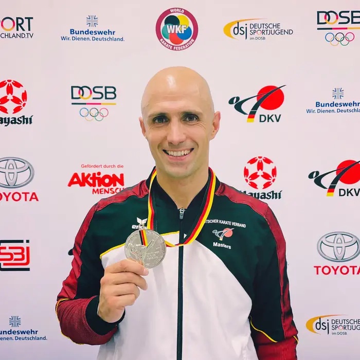 Thomas Lamm in Nationalmannschaftsjacke mit Silbermedaille vor einer Sponsorenwand